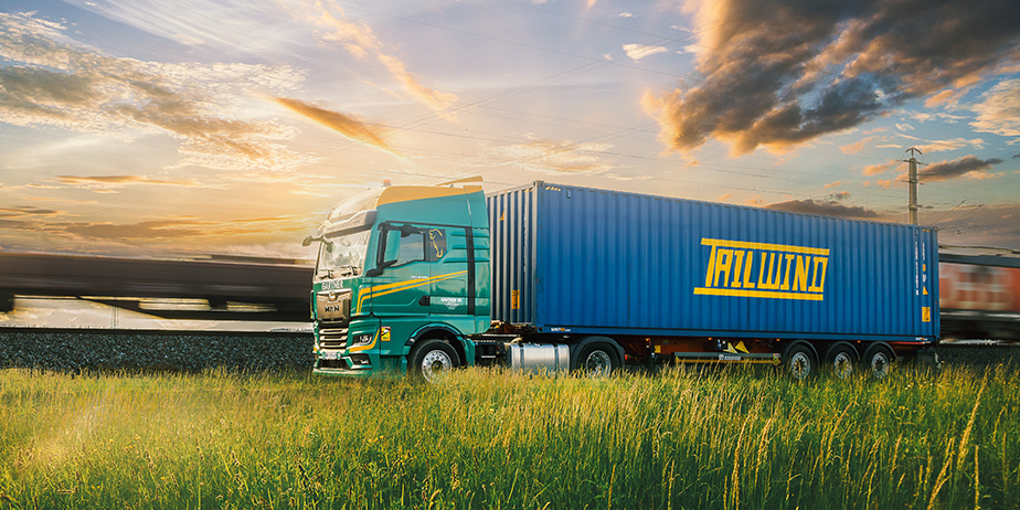 Blauer Schiffscontainer von TAILWIND auf einem GARTNER LKW in grüner Landschaft im Sonnenuntergang – GARTNER Transporte als Teil der TAILWIND Value Chain für punktgenaue Straßenlogistik in Europa.