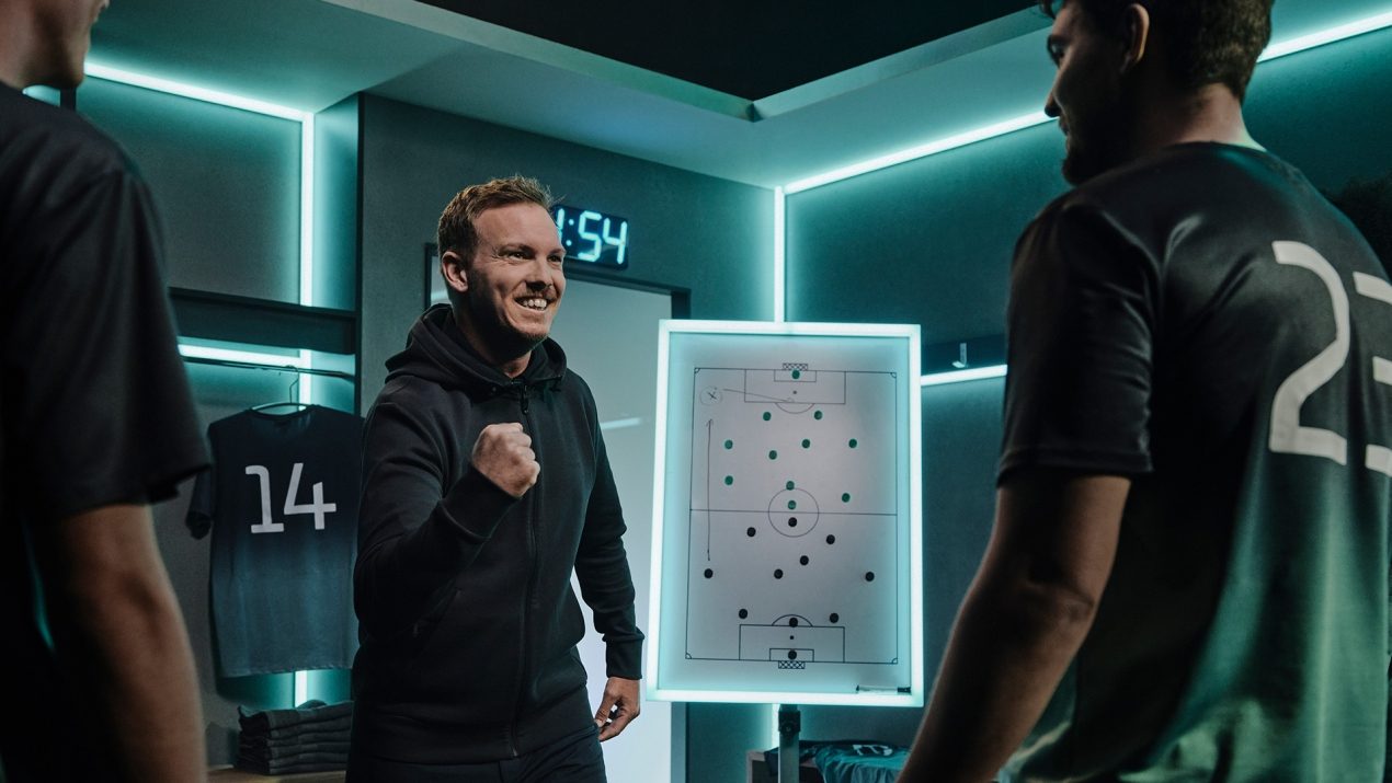 Julian Nagelsmann lächelt in einer futuristischen Umkleidekabine und zeigt die Faust, während er vor einem digitalen Taktikboard steht und mit zwei Fußballspielern spricht.