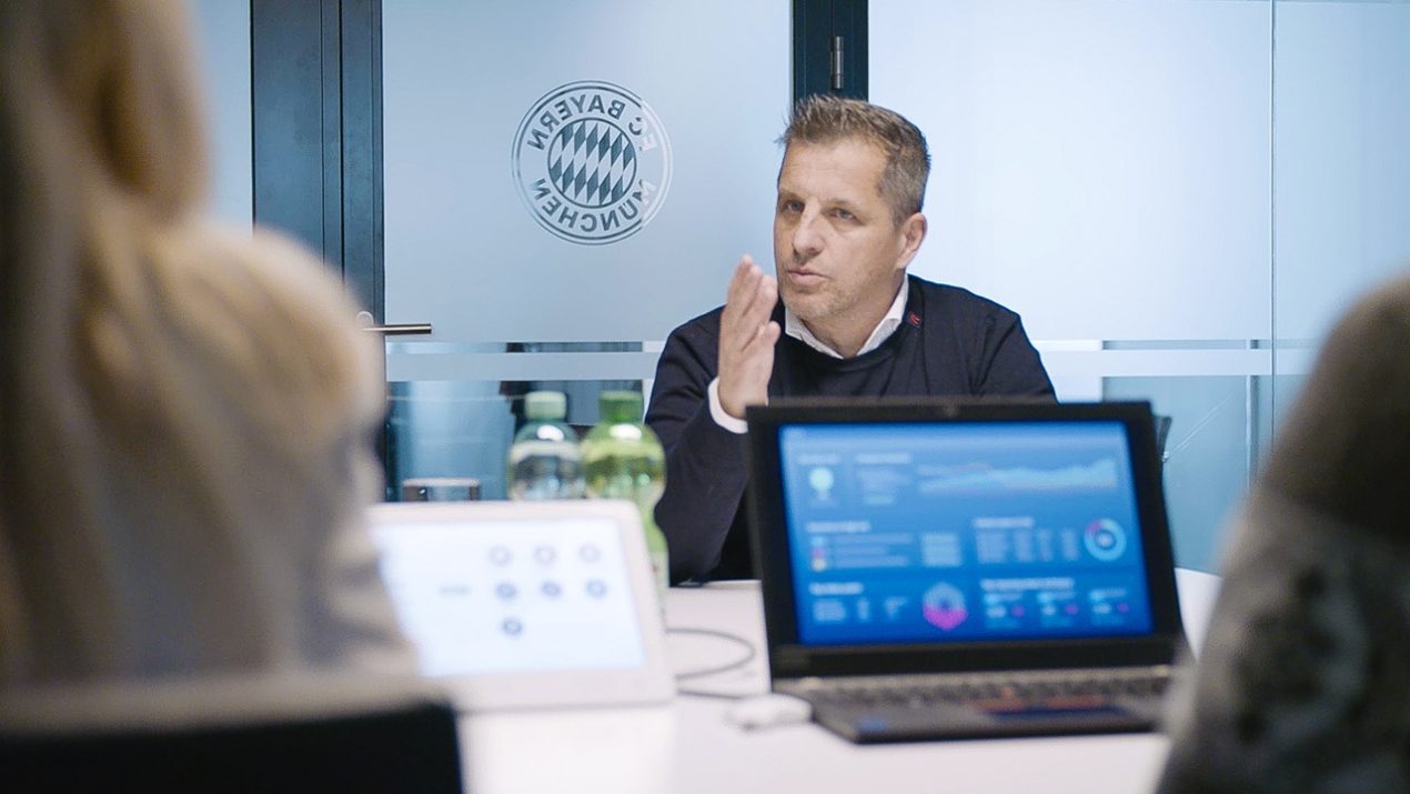 Das Bild zeigt ein Meeting. Im Hintergrund ist eine Tür mit dem Logo des FC Bayern München zu sehen. Vor der Tür sitzt ein weißer Mann mittleren Alters. Im Vordergrund sieht man den Rücken einer Frau mit langen blonden Haaren und rechts davon die Schulter einer weiteren Person. Es ist zu erkennen, dass die Personen sich unterhalten, da ein Laptop auf dem Tisch steht, der Diagramme zeigt.