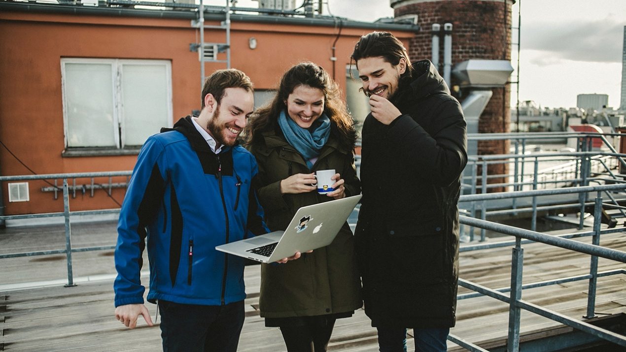 Drei Personen stehen zusammen und schauen auf einen Laptop und lächeln