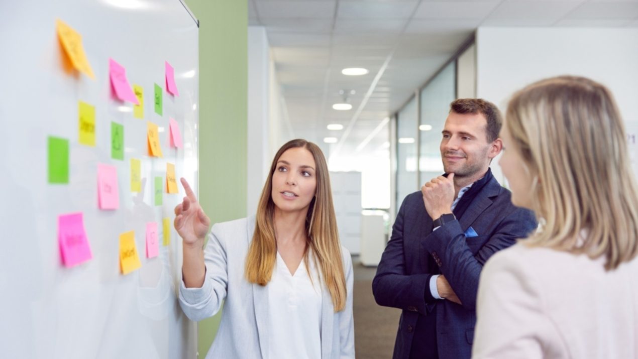 Drei Mitarbeiter stehen an einem Whiteboard, das mit Post-Its beklebt ist.