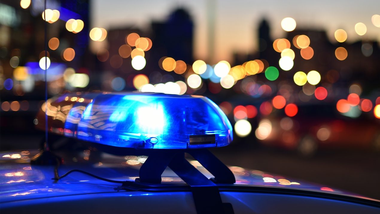 Close-up of a blue flashing light (emergency light) on the roof of an emergency vehicle (police or public order service) at dusk. In the blurry background, the lights and silhouette of a nighttime city with blurred, colorful bokeh effects from traffic are visible.