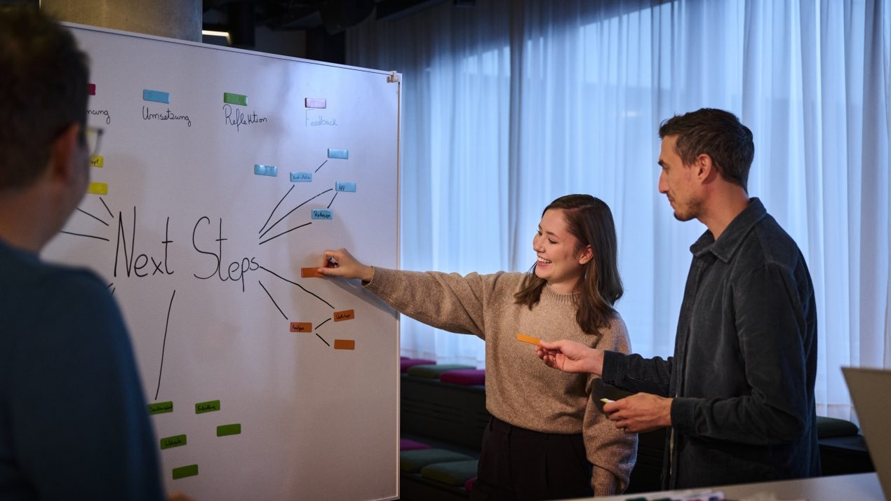 Drei Personen in einem Büroraum stehen vor einem Whiteboard mit der Aufschrift „Next Steps“ und bunten Haftnotizen.