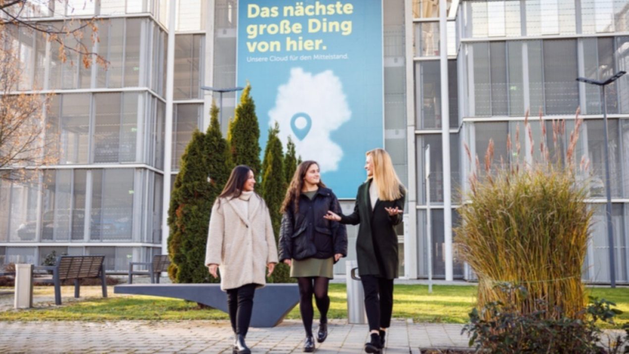 Drei Mitarbeiter laufen außen an einem Standort und sprechen miteinander. Im Hintergrund ist eine STACKIT Werbung zu sehen.