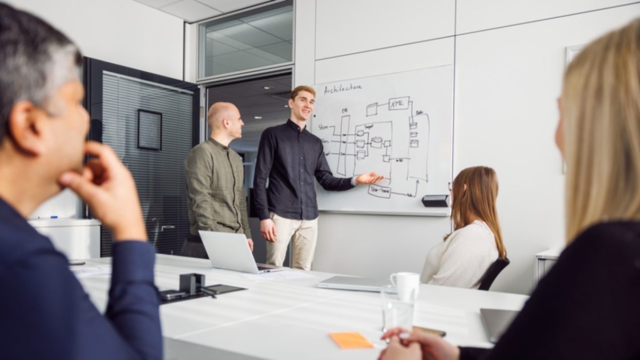 Insgesamt 5 Mitarbeiter der SIT sitzen in einem Besprechungsraum zusammen. Zwei Mitarbeiter stehen an einem beschriebenen Whiteboard und zeigen etwas.