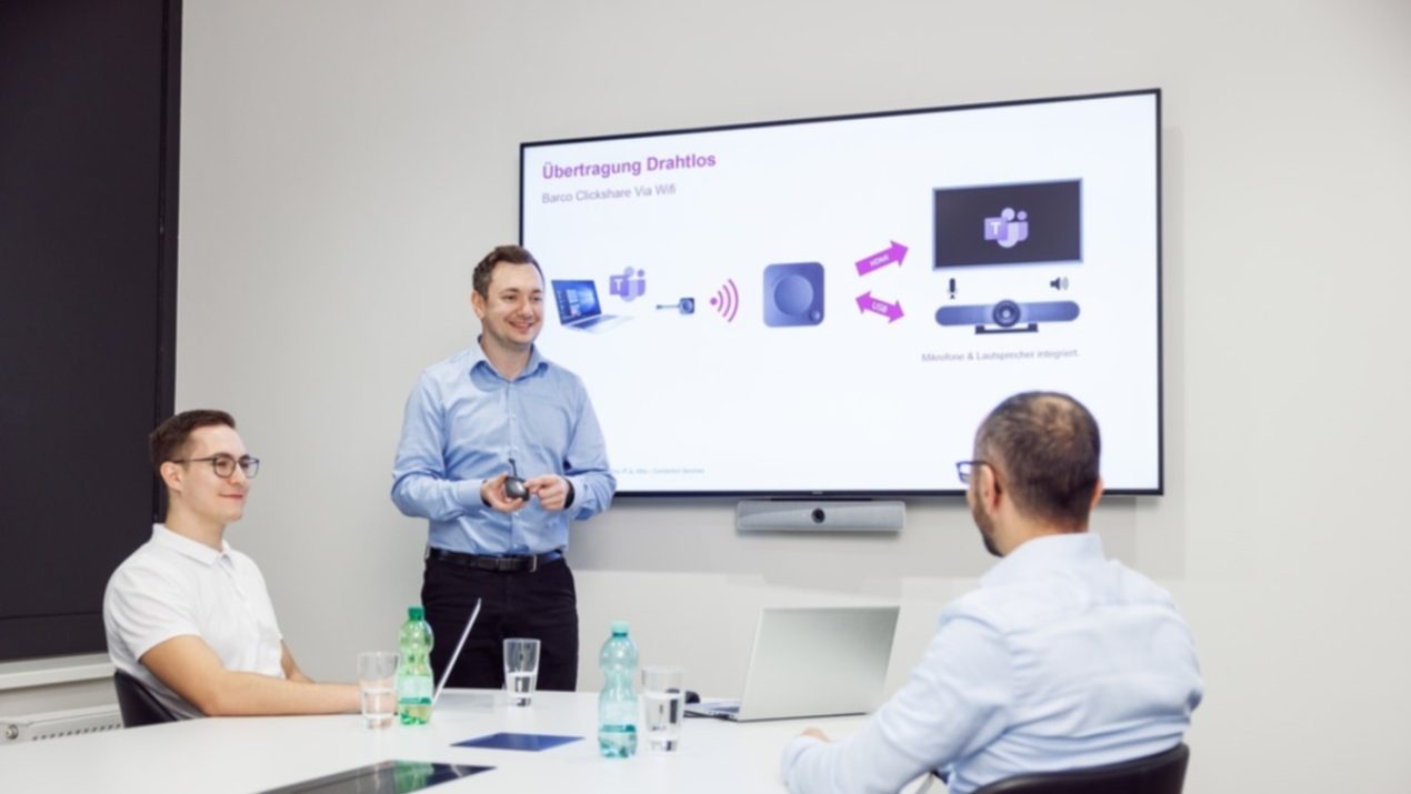 Drei Mitarbeiter sitzen in einem Konferenzraum zusammen. Ein Mitarbeiter steht vor dem Monitor und präsentiert etwas.