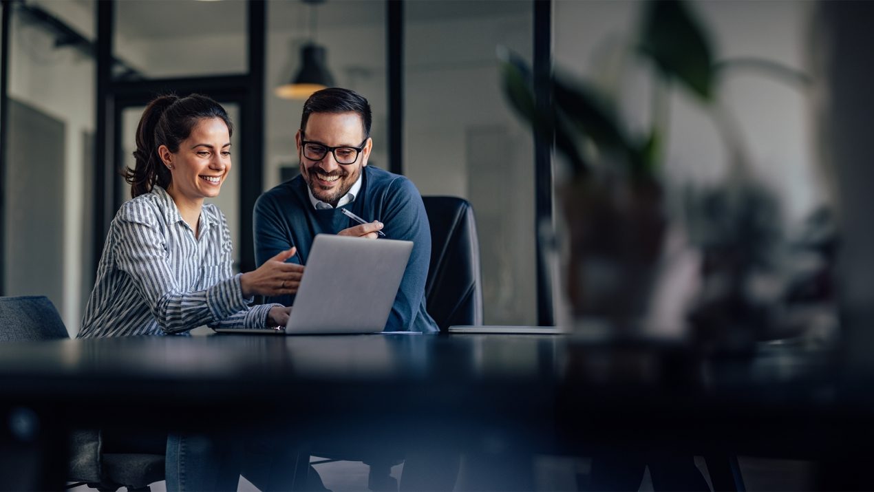 Auf dem Bild sind eine Frau und ein Mann zu sehen, die an einem Tisch sitzen. Sie lachen und schauen auf den vor ihnen stehenden grauen Laptop. Vermutlich befinden sie sich in einem Meeting. Im Vordergrund ist eine verschwommene grüne Pflanze zu sehen. Der Hintergrund zeigt einen verglasten Raum mit dunklen Rahmen.