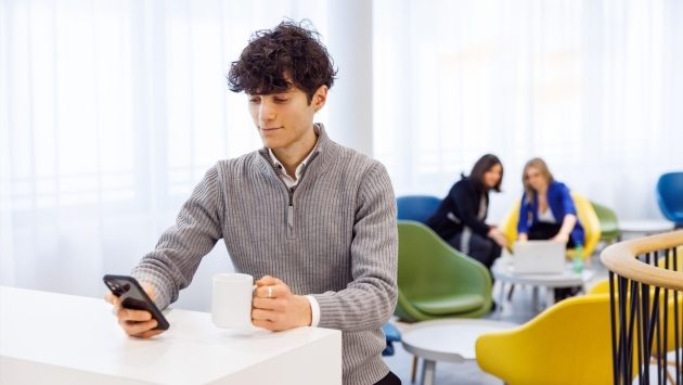 Ein Mitarbeiter sitzt mit Handy und Kaffeetasse an einem Tresen. Im Hintergrund sind zwei Mitarbeiterinnen an einem Laptop.
