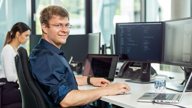 Ein Mitarbeiter sitzt an seinem Arbeitsplatz und lächelt in die Kamera. Auf seinen Monitoren sind Codes zu sehen.