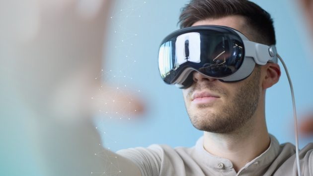 A light blue background can be seen. In the foreground is a young man in a beige T-shirt, short brown hair and VR glasses on his face. His right hand can be seen slightly blurred on the left side of the picture in the foreground, which he is stretching forward into the picture. A network is depicted in white above his hand.