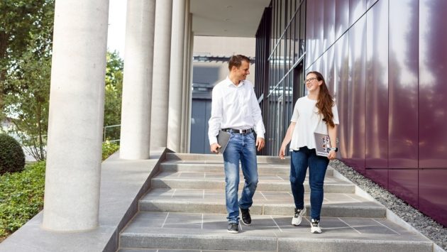 Zwei Mitarbeiter gehen die Treppe hinunter vor einem Bürogebäude und sprechen miteinander.