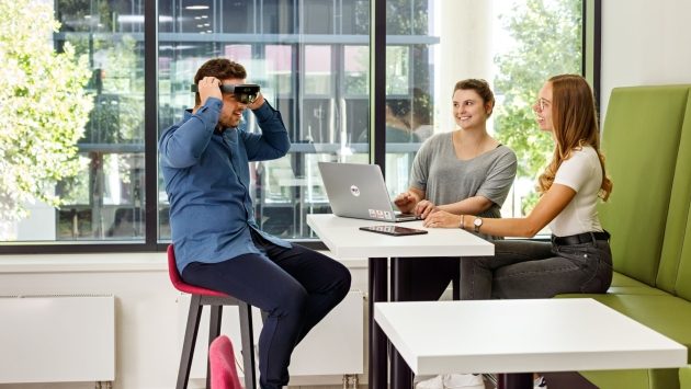Drei Mitarbeiter sitzen an einem Hochtisch. Ein Mitarbeiter setzt eine VR-Brille auf.