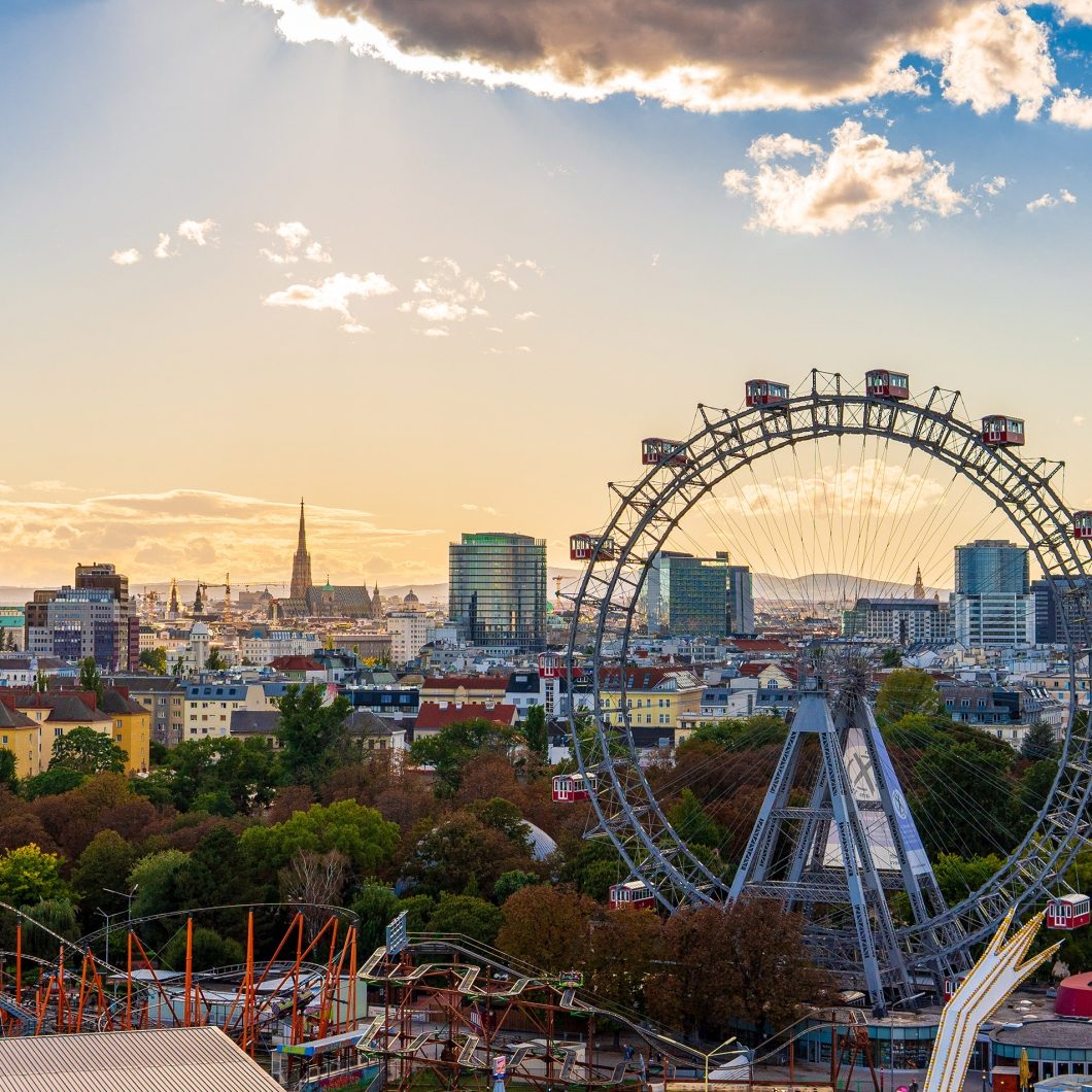 Ein Panoramablick auf die Wiener Skyline während der goldenen Stunde, dominiert vom historischen Wiener Riesenrad im Prater. Im Hintergrund ist der markante Turm des Stephansdoms inmitten der Stadtlandschaft zu sehen, während Sonnenstrahlen durch eine dramatische Wolkendecke am Abendhimmel brechen.