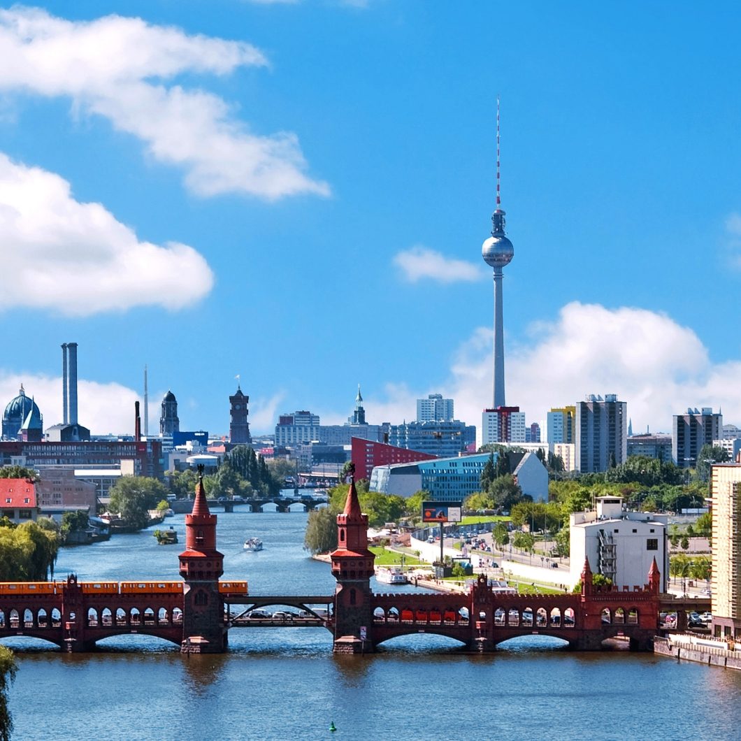 Panorama-Blick auf die Berliner Skyline mit der Oberbaumbrücke über der Spree und dem Berliner Fernsehturm im Hintergrund unter einem blauen Wolkenhimmel. Eine gelbe U-Bahn überquert gerade die Brücke.