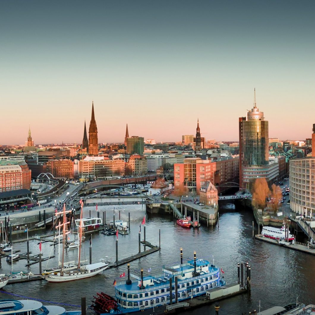 Panoramic view of Hamburg harbor at sunset featuring the Elbphilharmonie building on the right, boats docked in the waterway, and the city skyline in the background.