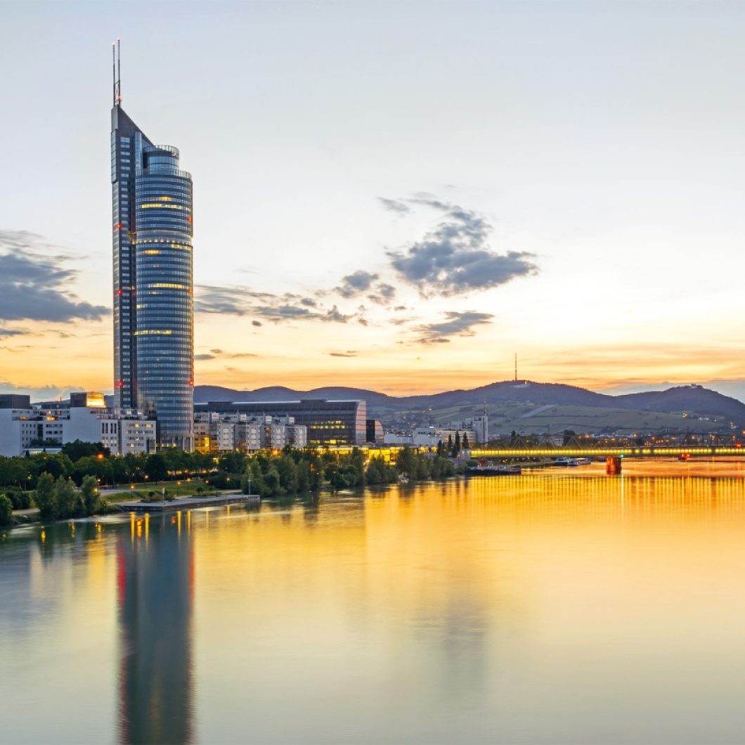 Millennium Tower skyscraper in Vienna, Austria, overlooking the Danube River during a golden sunset with mountain silhouettes in the background.