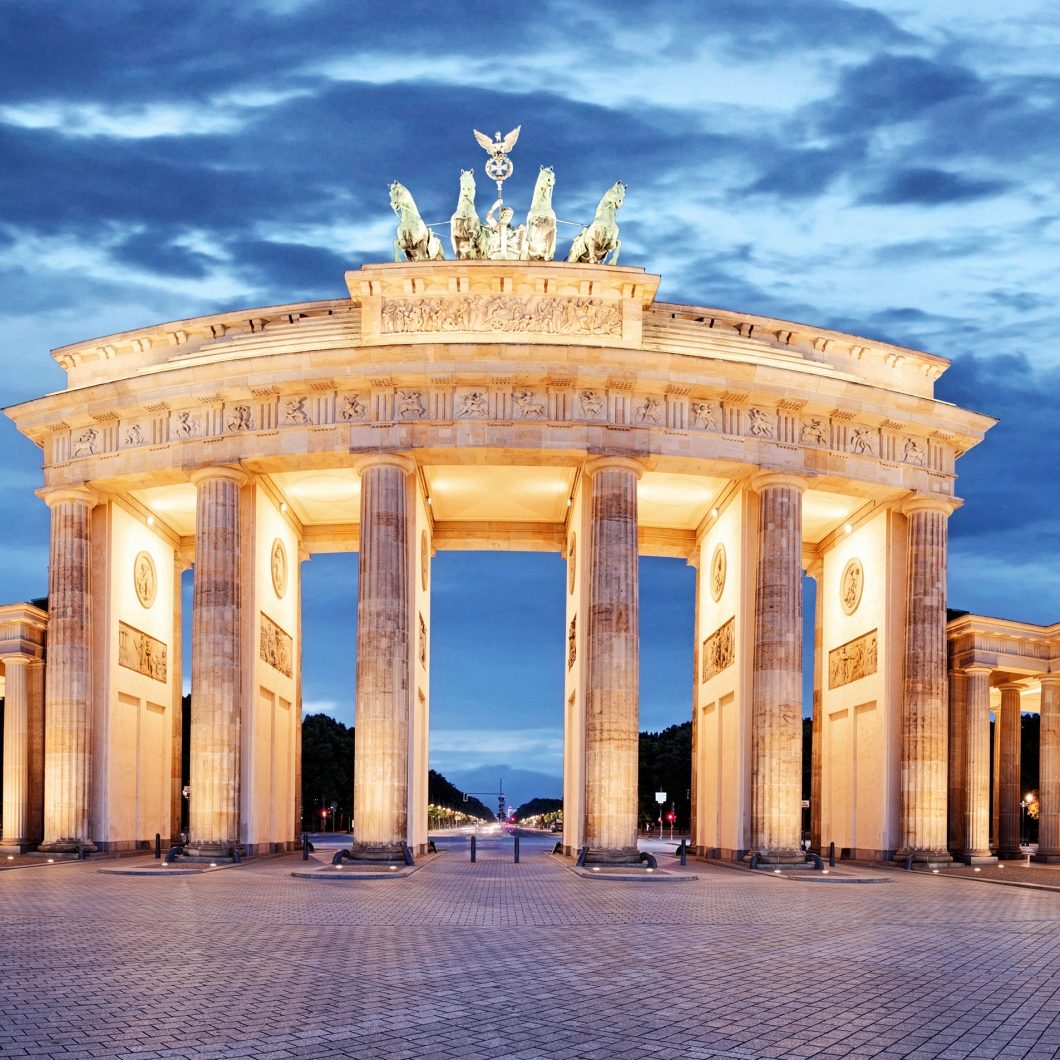 The Brandenburg Gate in Berlin at dusk, illuminated and framed by dark clouds. The night view emphasizes the imposing column architecture and the Quadriga on top.