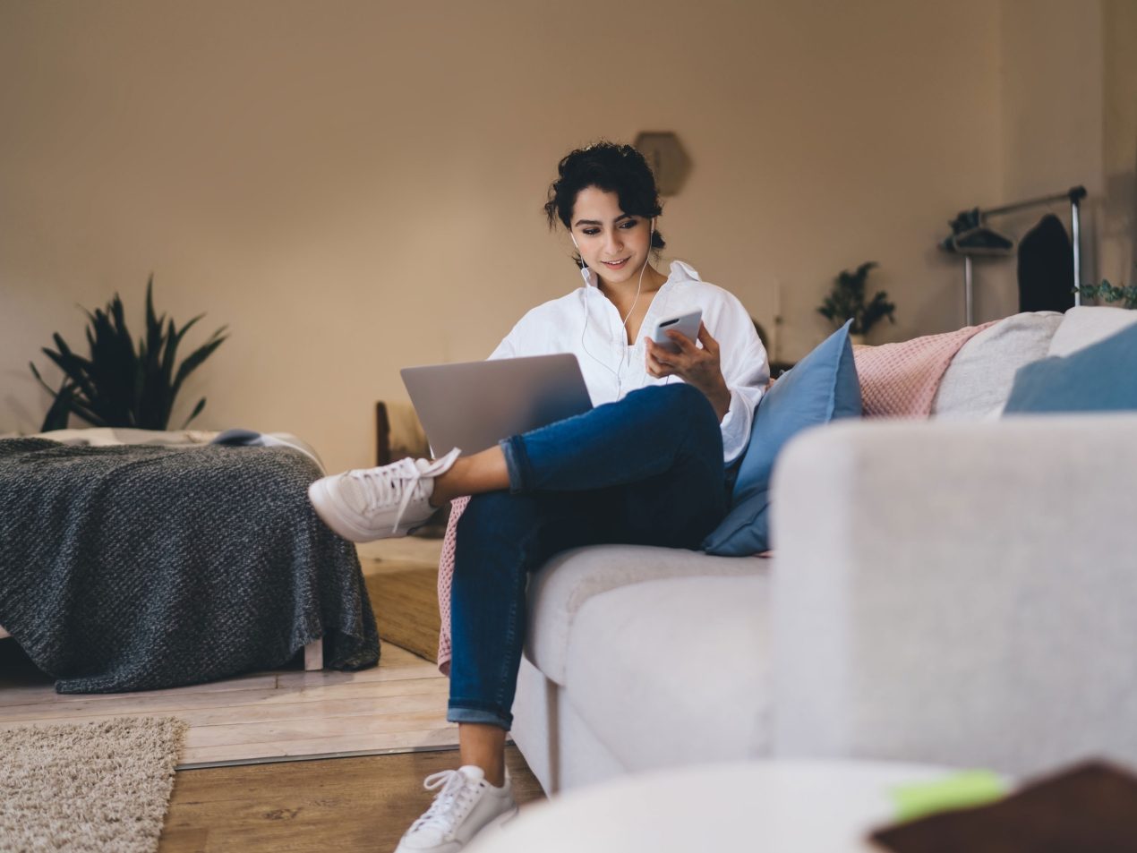 Young woman sitting comfortably on the sofa in her living room, wearing earphones and working simultaneously with a laptop and smartphone. Symbolizes home office, flexible working, and remote learning.