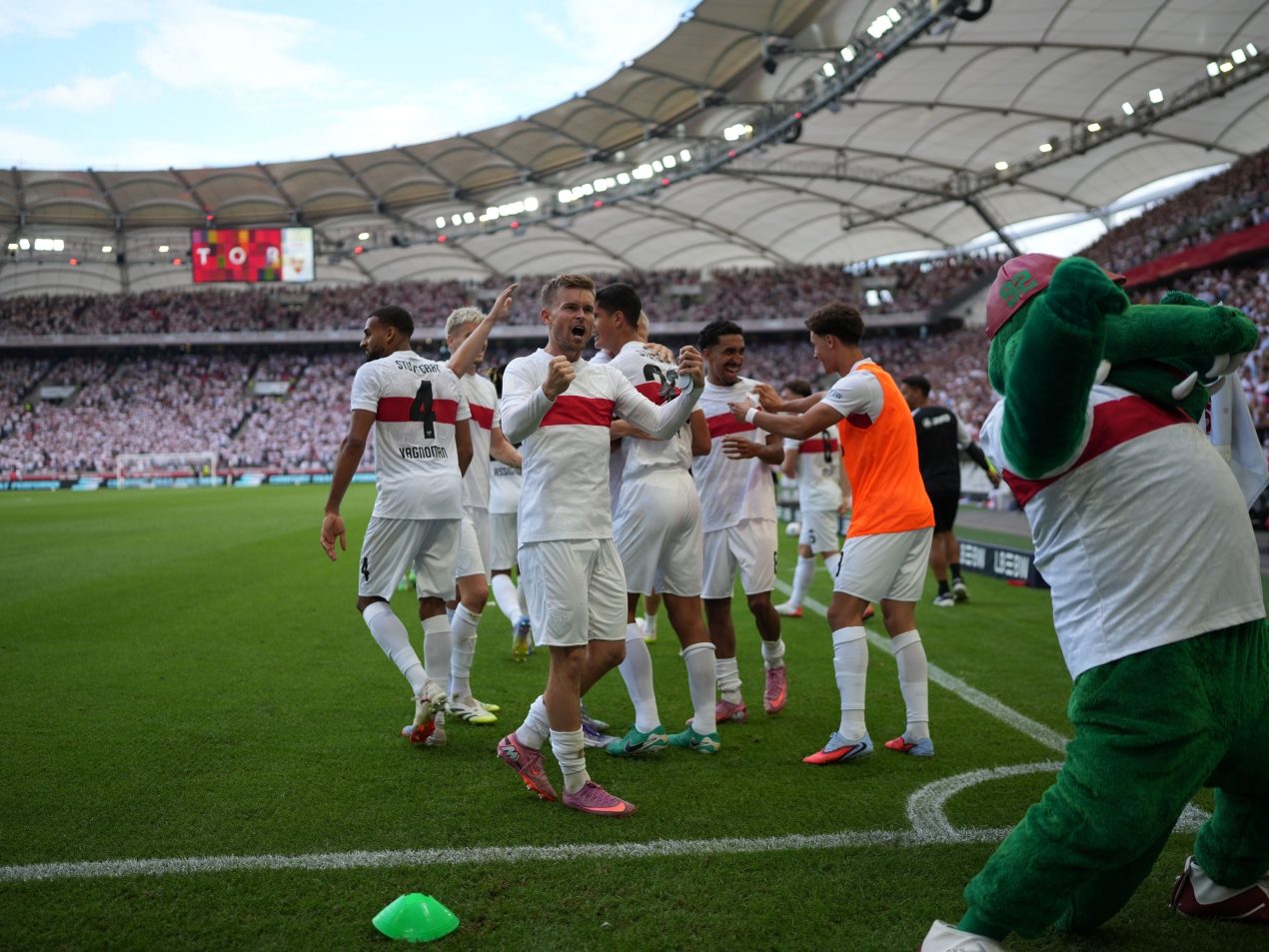 Jubelnde VfB-Spieler im Kreis auf dem Rasen. Das Maskottchen Fritzle steht im Vordergrund und teilt die Freude mit der Mannschaft.