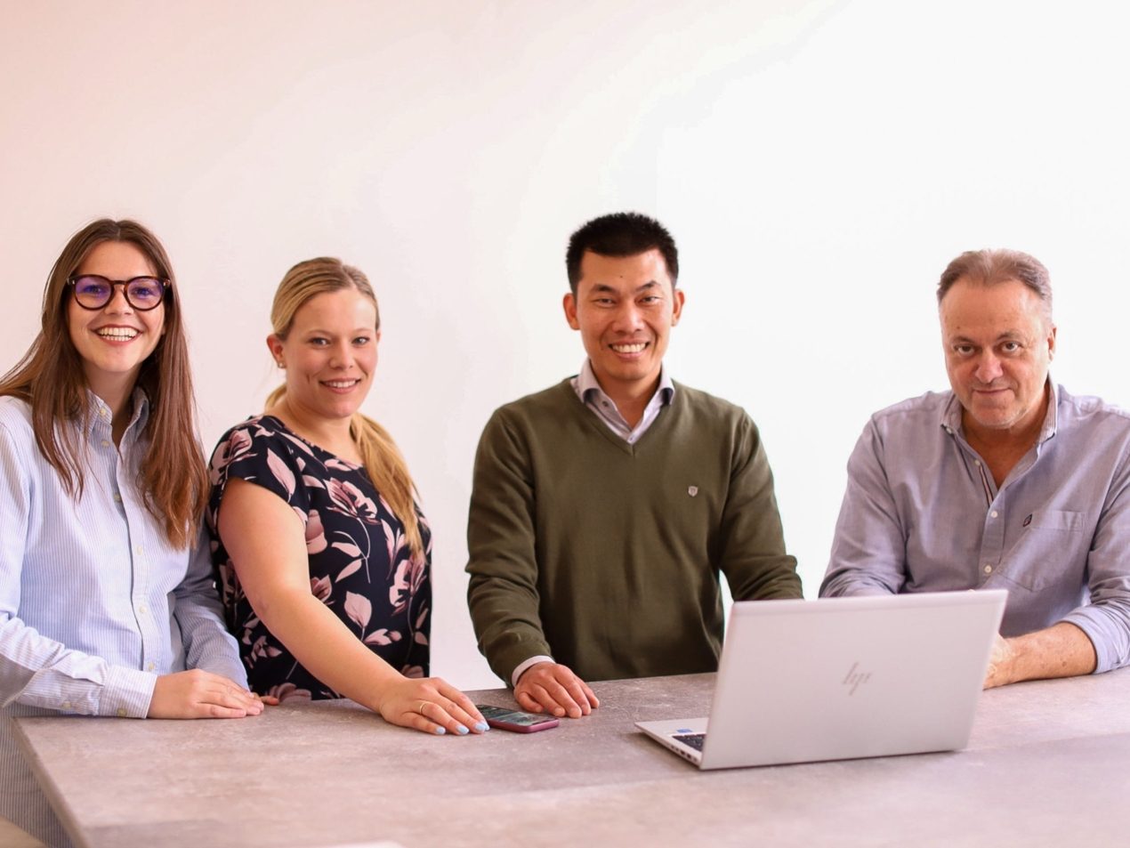 Vier Mitarbeiter stehen an einem Tisch, schauen in die Kamera. Vor ihnen steht ein Laptop.