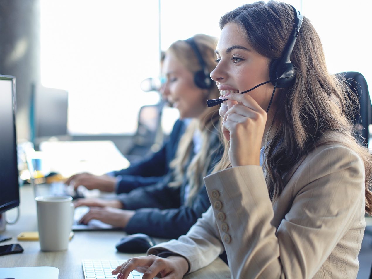 Seitenprofil einer lächelnden Frau im hellen Blazer. Sie trägt ein Headset, berührt das Mikrofon und arbeitet am Computer. Im unscharfen Hintergrund sitzen weitere Kollegen an ihren Bildschirmen.