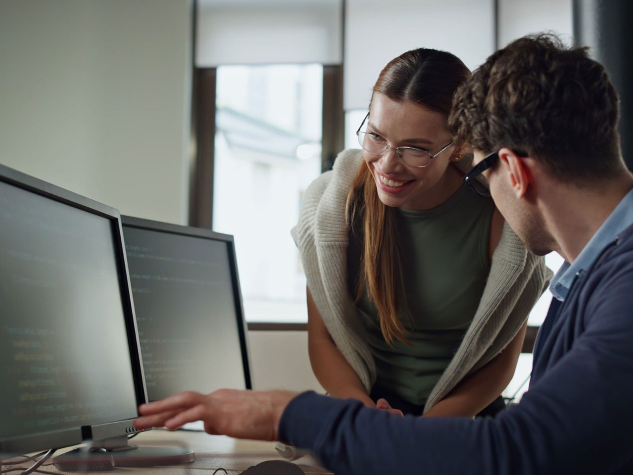 Eine Frau und ein Mann im Büro blicken gemeinsam lächelnd auf einen Bildschirm mit Programmcode, auf den der Mann zeigt.