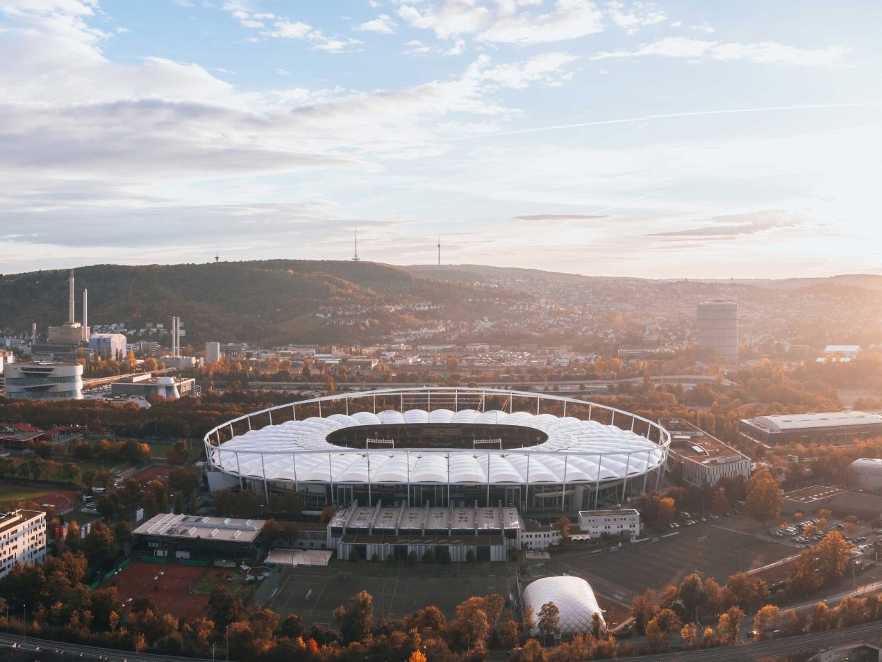 Optimierter Alt-Text:  Panoramische Luftaufnahme der MHPArena (ehemals Mercedes-Benz Arena/Neckarstadion) in Stuttgart, der Heimstätte des VfB Stuttgart, eingebettet in den NeckarPark. Das moderne Stadion mit seiner markanten weißen Dachkonstruktion und dem Ring steht im Zentrum des Bildes, umgeben von der städtischen Bebauung Stuttgarts und bewaldeten Hügeln im Hintergrund. Die Szene wird von warmem Sonnenlicht durchflutet, das am späten Nachmittag oder frühen Morgen über der Stadt scheint und eine herbstliche Stimmung erzeugt. Ideal zur Bebilderung von Sport, Events, Architektur oder der Stadt Stuttgart.