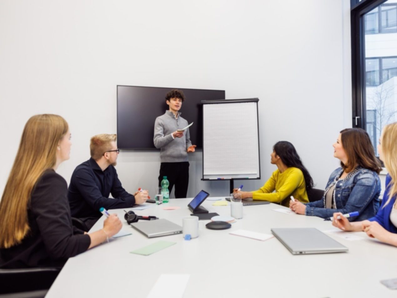 Ein Mitarbeiter steht vorne in einem Besprechungsraum an einem Flipchart. 5 weitere Mitarbeiter hören ihm zu.