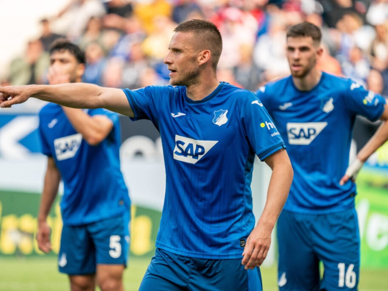 Fußballspieler der TSG Hoffenheim (blaues Trikot, SAP-Sponsor) gibt Anweisungen auf dem Spielfeld. Er zeigt mit dem Finger und blickt über das Spielfeld, während Teamkollegen im Hintergrund warten.