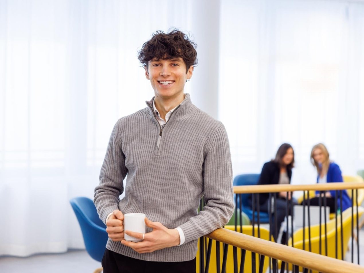 Ein Mitarbeiter steht an einem Geländer mit Kaffeetasse in der Hand. Er lächtelt in die Kamera. Im Hintergrund sind zwei weitere Mitarbeiterinnen zu sehen.