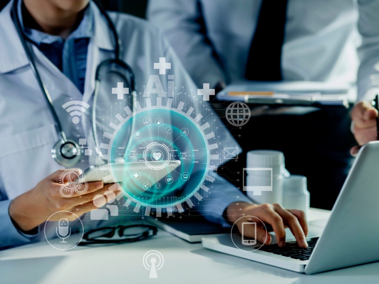 A doctor or medical professional in a white coat with a stethoscope types on a laptop while holding a smartphone. Above the laptop, a holographic projection of a blue circle with a heart symbol in the center and many smaller icons representing digital health services (e.g., Wi-Fi, communication, medication) hovers. Another person in a shirt is in the background. The image symbolizes e-health, telemedicine, and digital transformation in healthcare.