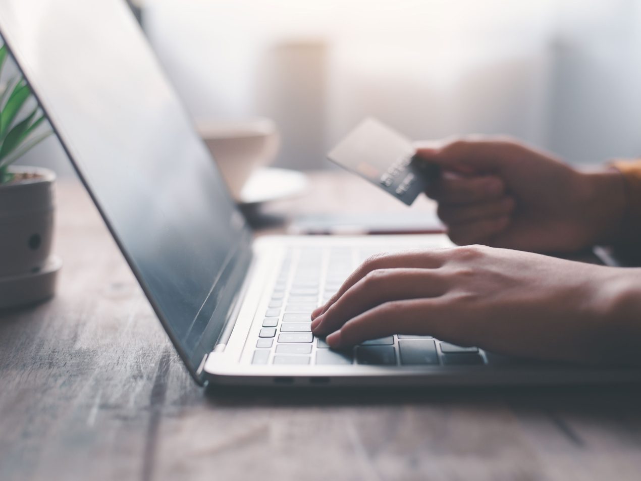 Nahaufnahme von Händen, die auf einem Laptop tippen und eine Kreditkarte halten. Symbolisiert Online-Shopping, sichere Zahlung oder Mobile Banking auf einem Holztisch.