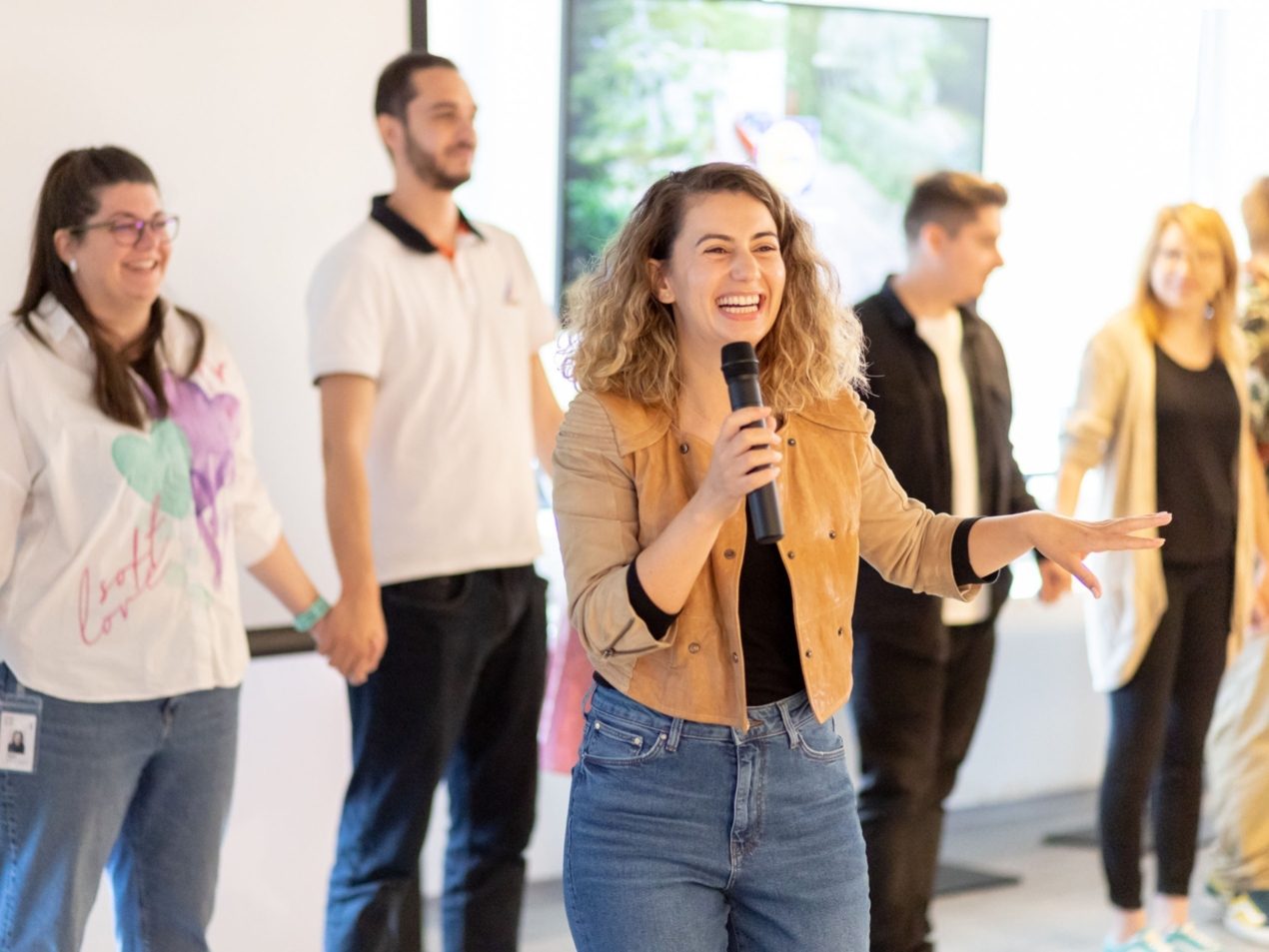 Eine Mitarbeitern steht vor ihren Kolleginnen und Kollegen mit einem Mikrophon. Alle lachen und haben Freude.