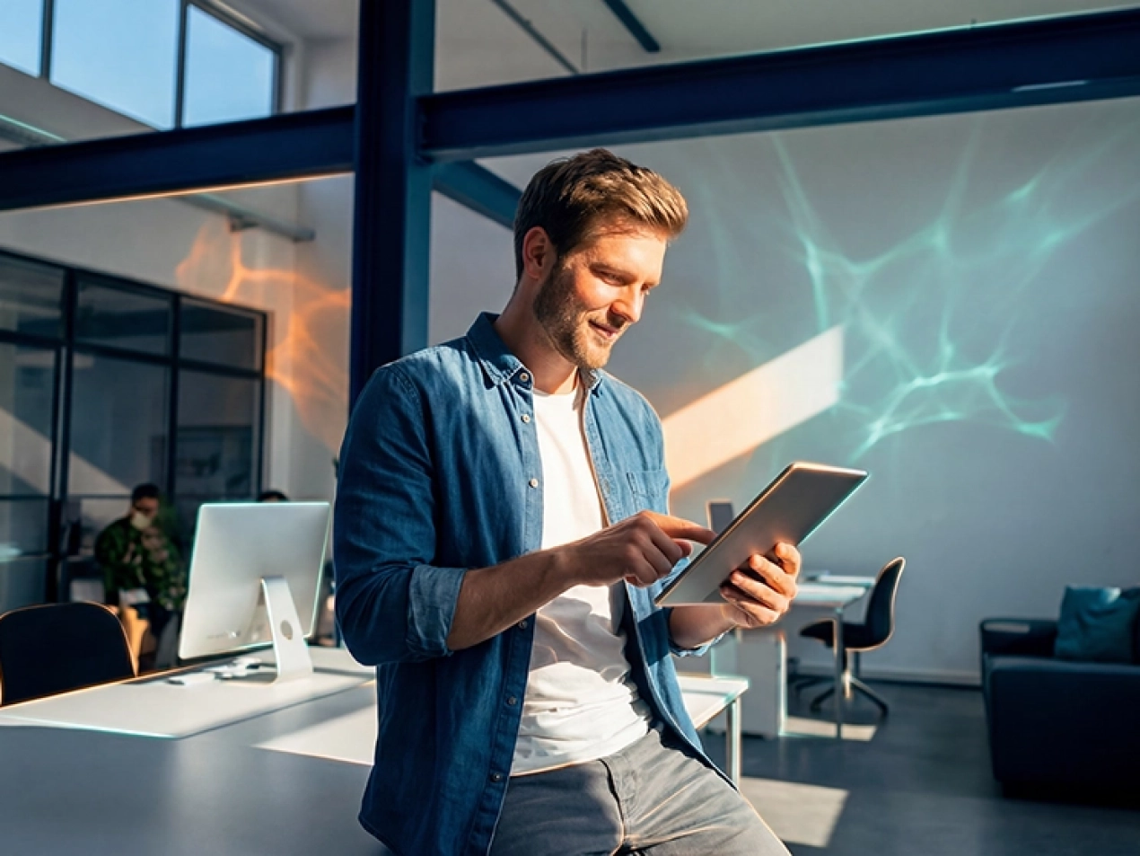 Ein lächelnder Mann in einem blauen Hemd steht in einem hellen, modernen Büro und bedient ein Tablet, während Lichtreflexionen im Hintergrund eine dynamische Atmosphäre schaffen.