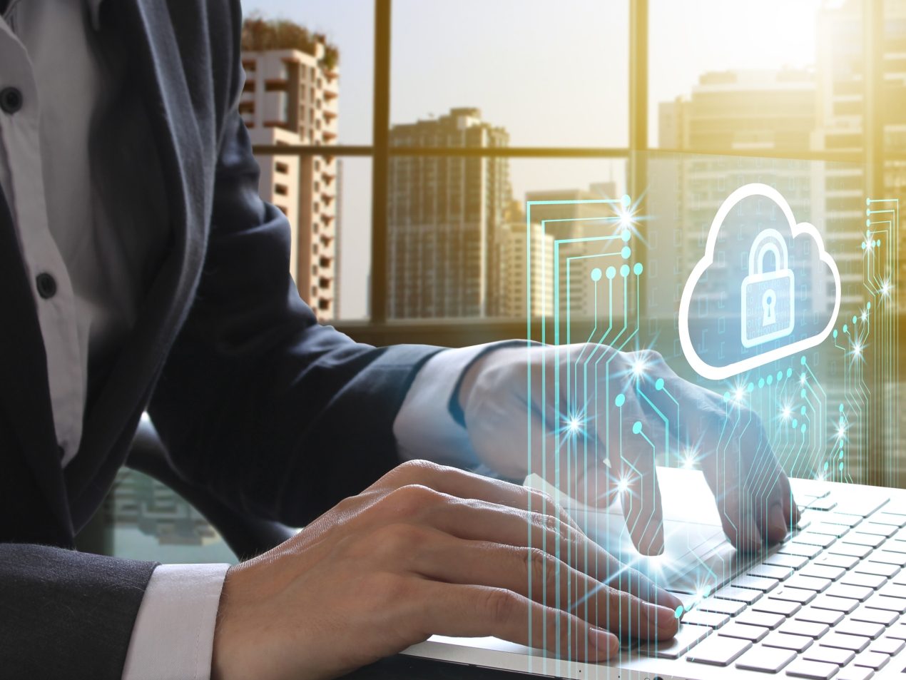 A business person in a suit types on a laptop keyboard in front of a large window with a city view in the sunlight. A futuristic, glowing blue projection of a cloud with a padlock (symbolizing cloud security/data protection) and connected circuits is superimposed above the laptop.