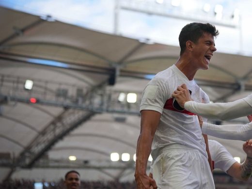 Jubilant VfB Stuttgart football players (in white home jerseys) celebrating a successful goal inside the stadium. One player shouts in joy while being embraced by teammates.