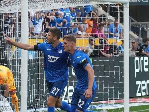 TSG Hoffenheim soccer players (in blue jerseys with numbers 27 and 22) celebrate a goal in front of the net. The opposing goalkeeper is on the ground, and fans cheer in the stands. A "Zero Waste" banner by PreZero is visible in the stadium.