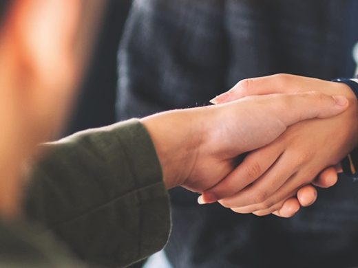 Nahaufnahme von zwei Personen, die sich die Hände schütteln, was eine Geschäftsvereinbarung oder Partnerschaft symbolisiert. Eine Person trägt ein olivgrünes Oberteil, die andere eine Jacke mit gestreiften Manschetten. Der Hintergrund ist verschwommen, mit einem Tisch oder Schreibtisch, der auf ein Büro- oder Besprechungsraum-Setting hindeutet.