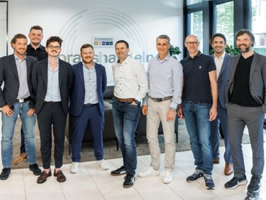 Gruppe von neun professionellen Männern, die in einer modernen Büro-Lobby mit großen Fenstern und einem Schwarz-Blockchain-Logo an der Wand stehen.