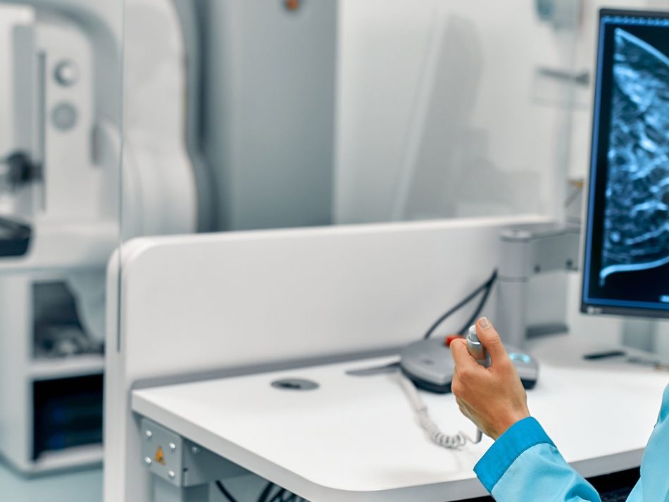 Medical professional examining mammogram images on a screen with a patient standing at the mammography machine.