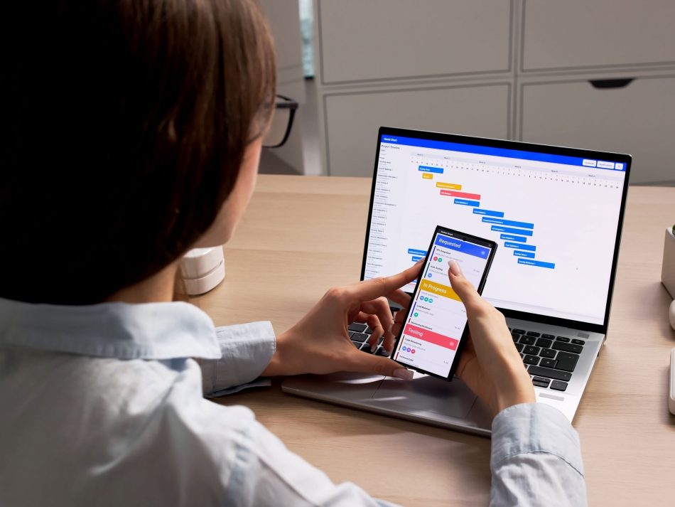 Person at a desk managing a Kanban task board on a smartphone alongside a project Gantt chart on a laptop.