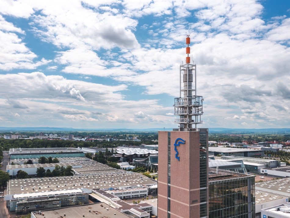 Aerial view of the Messeturm (Hermes Tower) on the grounds of the Deutsche Messe in Hannover under a cloudy sky. The brown-glass tower with the blue logo on its facade and the distinctive antenna structure rises above the flat halls of the exhibition center.