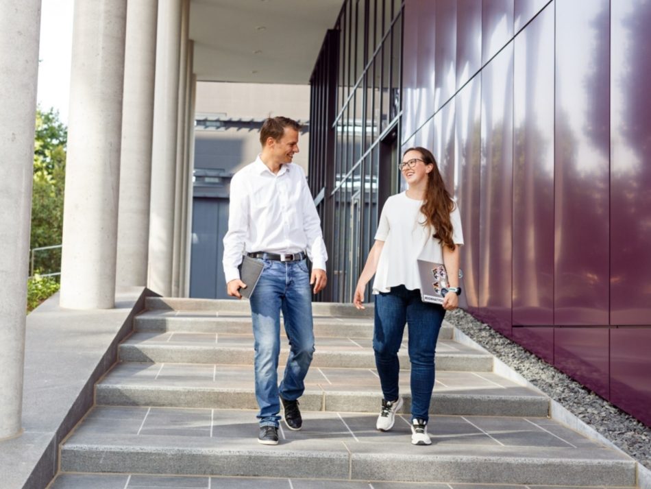 Zwei Mitarbeiter gehen die Treppe hinunter vor einem Bürogebäude und sprechen miteinander.