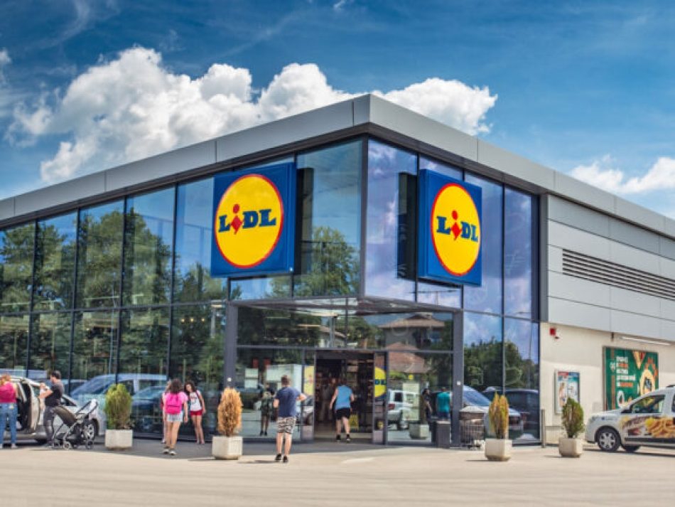 Außenansicht einer modernen Lidl-Filiale mit großflächiger Glasfassade und dem gelb-blauen Lidl-Logo, an einem sonnigen Tag. Das Bild zeigt den Lebensmitteleinzelhandel, das Lidl-Gebäude und Kunden beim Betreten und Verlassen des Supermarktes.