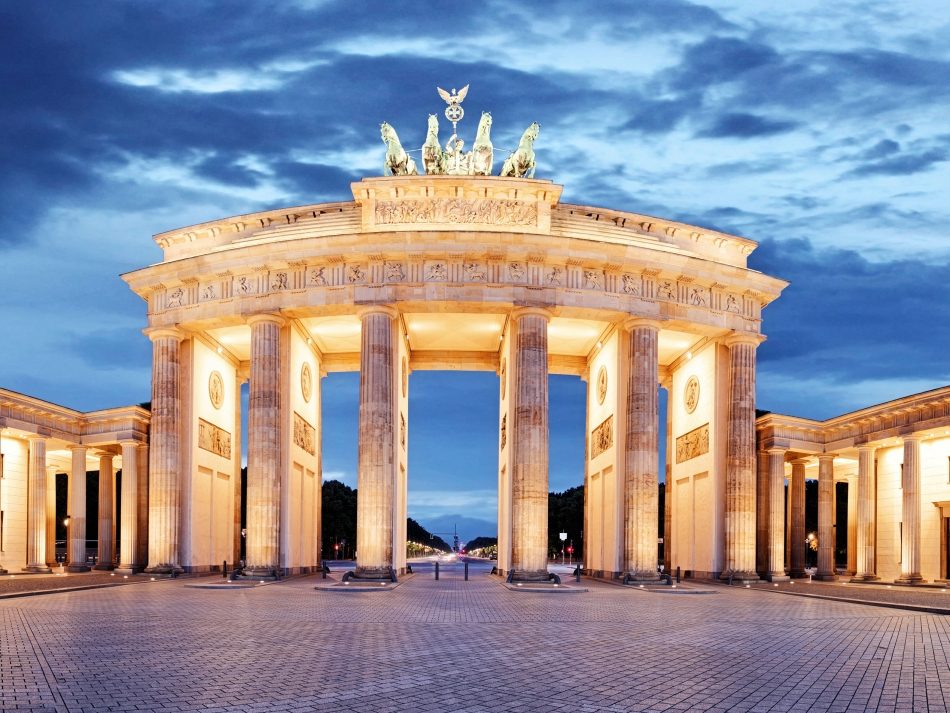 The Brandenburg Gate in Berlin at dusk, illuminated and framed by dark clouds. The night view emphasizes the imposing column architecture and the Quadriga on top.