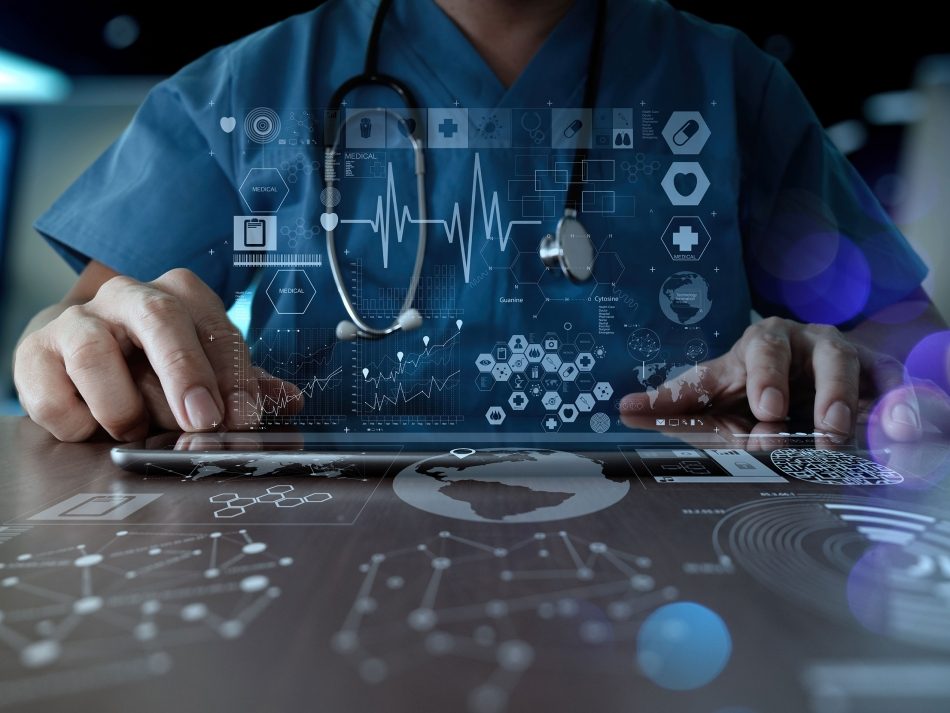 A doctor in blue scrubs and a stethoscope around his neck is sitting at a desk. He is tapping on a futuristic, transparent tablet that projects holographic medical symbols, data graphs, and a global map onto the table surface. The scene conveys a sense of digital healthcare, technology, and medical innovation.