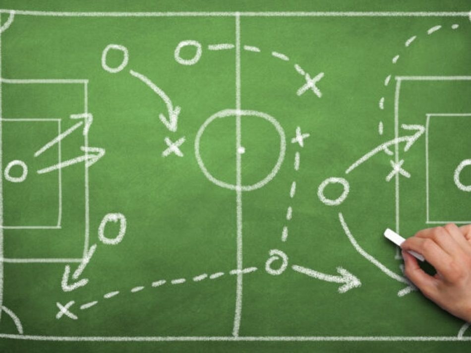 Eine Hand zeichnet mit Kreide auf einer grünen Tafel eine Spieltaktik oder Strategie auf einem Fußballfeld-Schema (Taktiktafel) ein. Das Bild symbolisiert strategische Planung, Taktikentwicklung, Geschäftsstrategie, Teamentwicklung und Coaching.