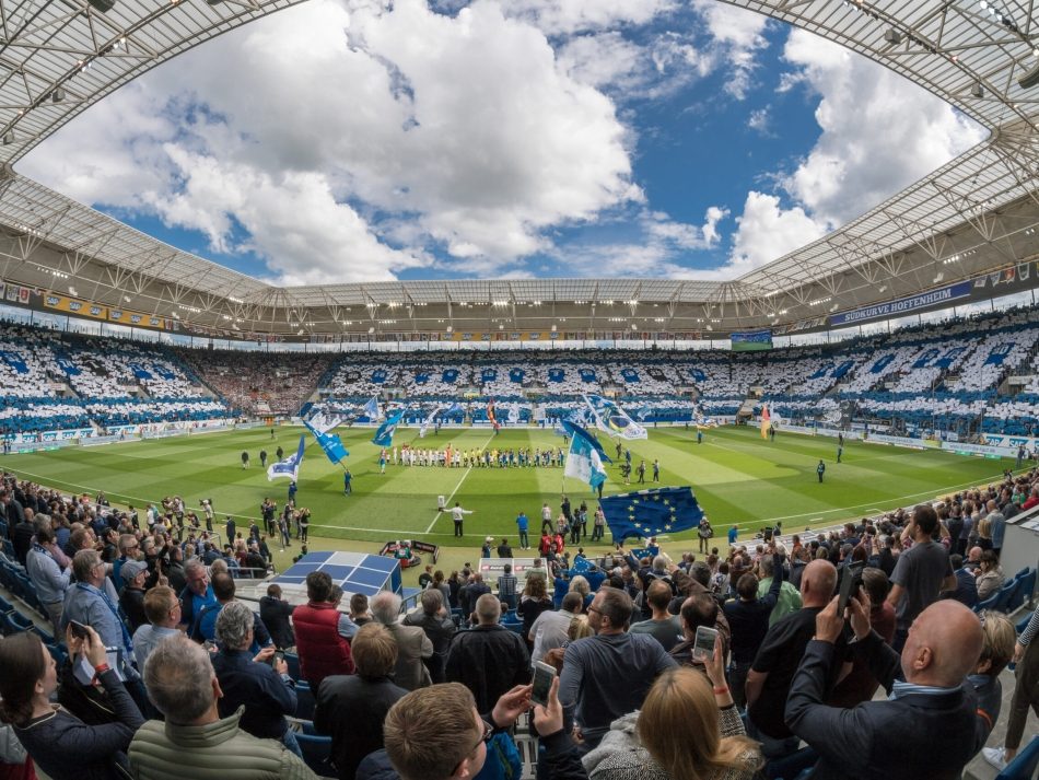 Weitwinkelaufnahme des Innenraums der PreZero Arena, des Stadions der TSG Hoffenheim. Die Tribünen sind gefüllt mit Fans, die eine blau-weiße Choreographie zeigen, während die Mannschaften auf das Spielfeld laufen.