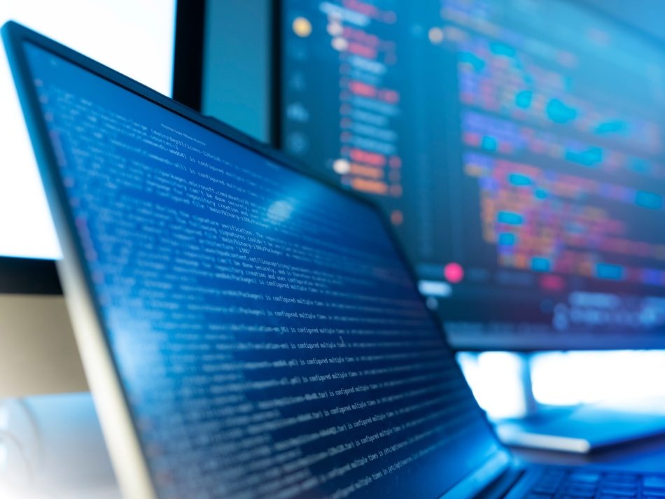 Nahaufnahme eines Laptop-Bildschirms mit blauem Programmiercode, im Hintergrund weitere Monitore mit Quellcode an einem modernen IT-Arbeitsplatz.