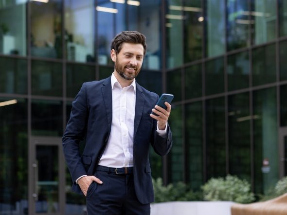 A smiling businessman in a dark blue suit stands in front of a modern office building with a glass facade, looking at his smartphone. This image was generated with AI.